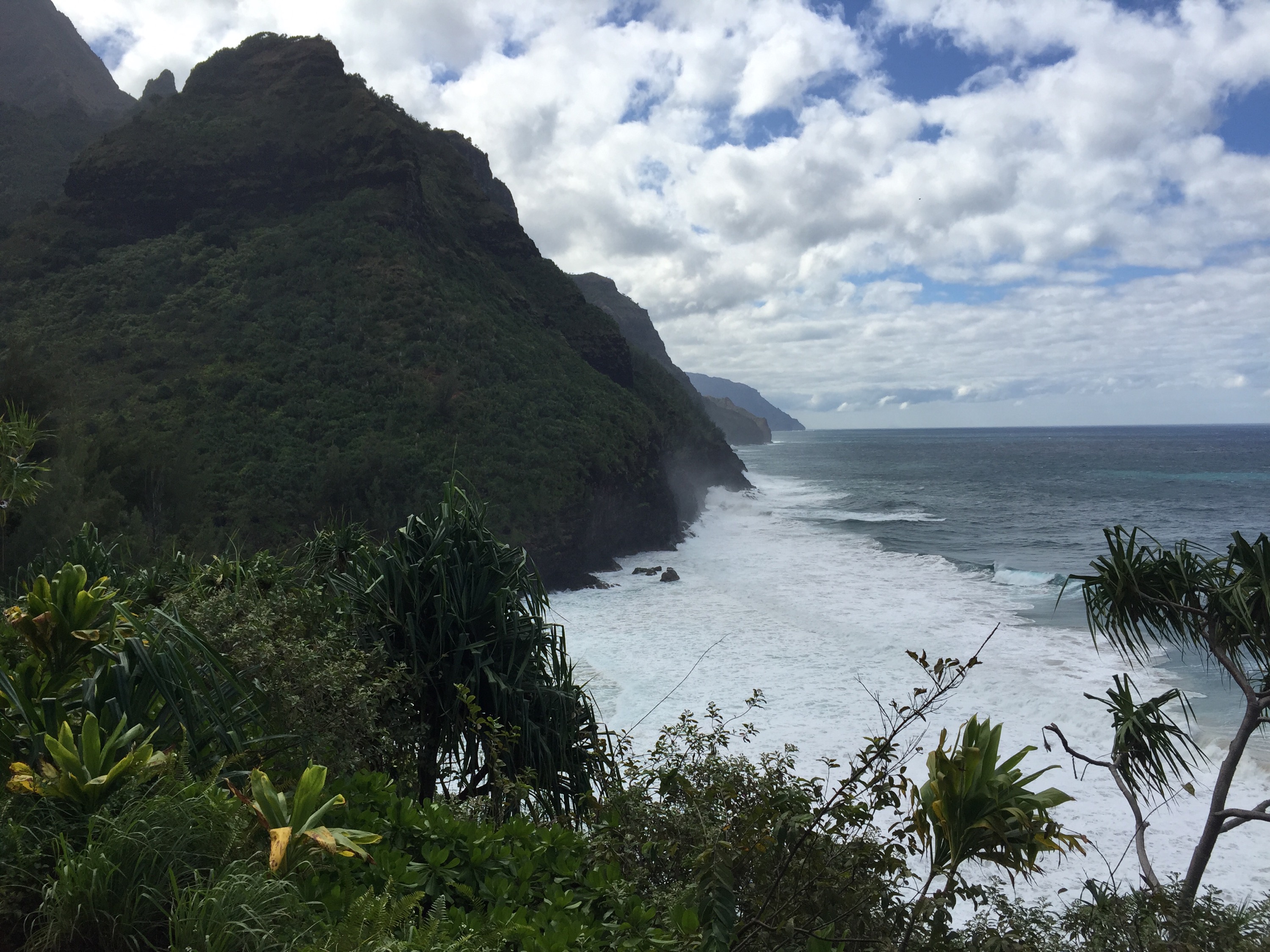 Gorgeous green foliage covers steep and tall green mountains which jut up from the sea. The sky is blue with clouds.