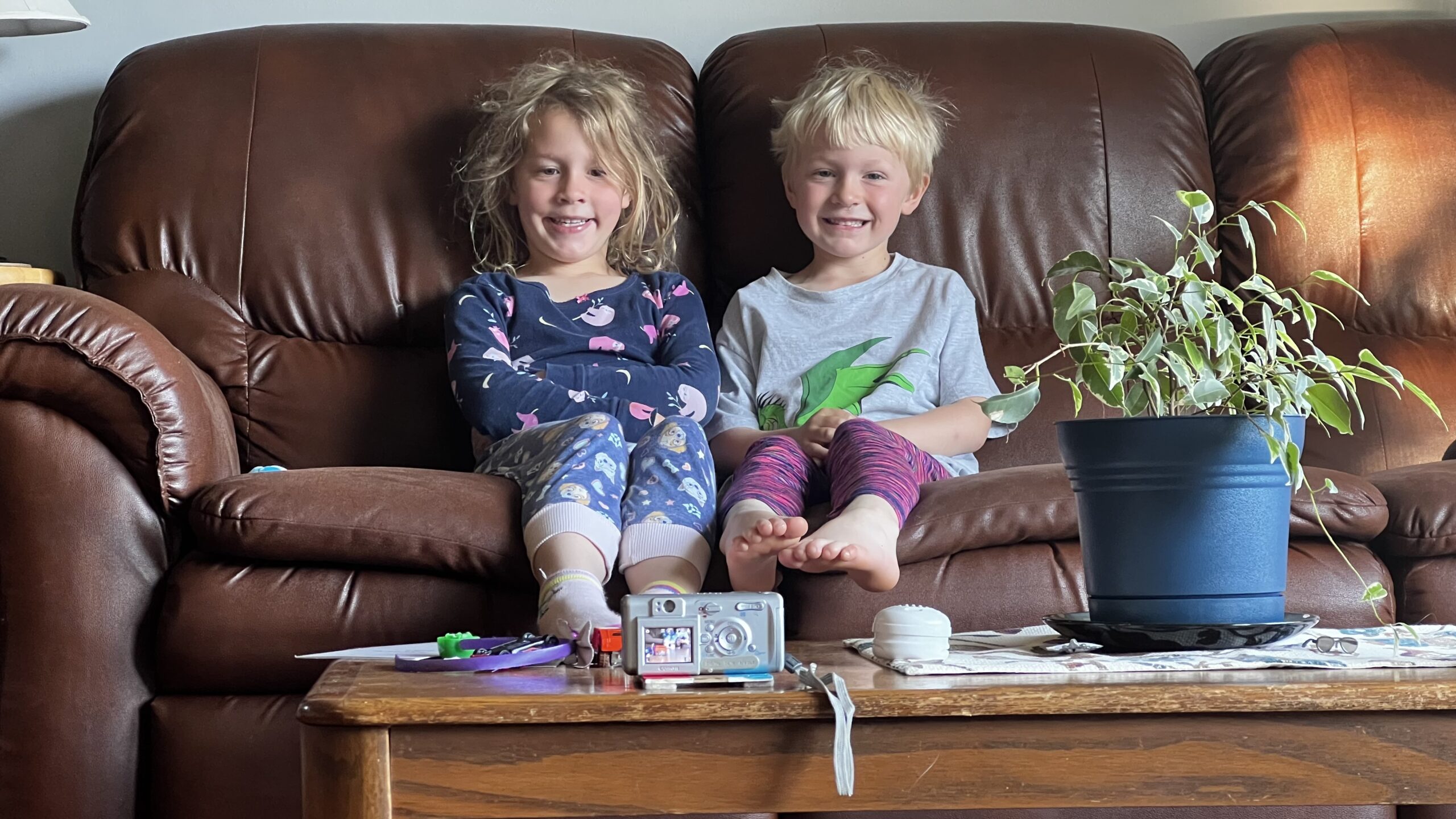 Two young children sit on a sofa with a digital camera set up on self timer mode takes their photo.